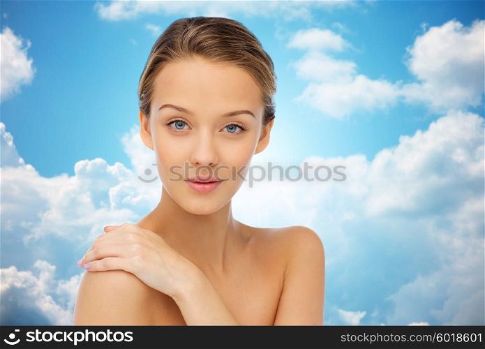 beauty, people, body care and health concept - smiling young woman face and hand on bare shoulder over blue sky and clouds background