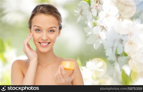 beauty, people, cosmetics, skincare and cosmetics concept - happy young woman applying cream to her face over summer green natural background with cherry blossom