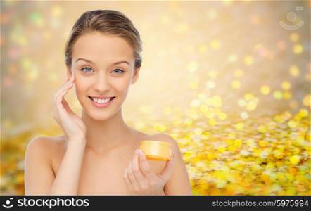beauty, people, cosmetics, skincare and cosmetics concept - happy young woman applying cream to her face over golden glitter or holidays lights background