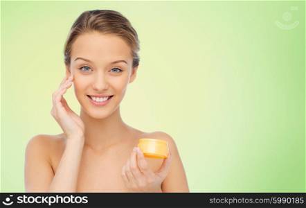 beauty, people, cosmetics, skincare and cosmetics concept - happy young woman applying cream to her face over green natural background