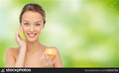 beauty, people, cosmetics, skincare and cosmetics concept - happy young woman applying cream to her face over green natural background