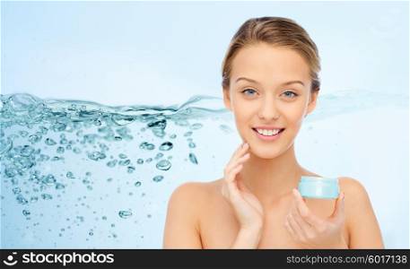 beauty, people, cosmetics, skincare and cosmetics concept - happy young woman applying cream to her face over water splash on blue background