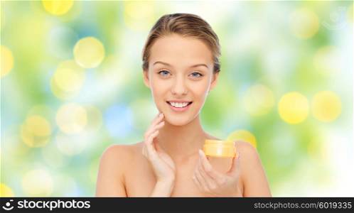 beauty, people, cosmetics, skincare and cosmetics concept - happy young woman applying cream to her face over green lights background