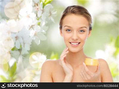 beauty, people, cosmetics, skincare and cosmetics concept - happy young woman applying cream to her face over green natural background with cherry blossom