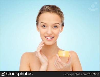 beauty, people, cosmetics, skincare and cosmetics concept - happy young woman applying cream to her face over blue background