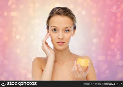 beauty, people, cosmetics, skincare and cosmetics concept - young woman applying cream to her face over rose quartz and serenity lights background