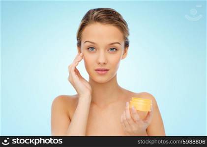beauty, people, cosmetics, skincare and cosmetics concept - young woman applying cream to her face over blue background
