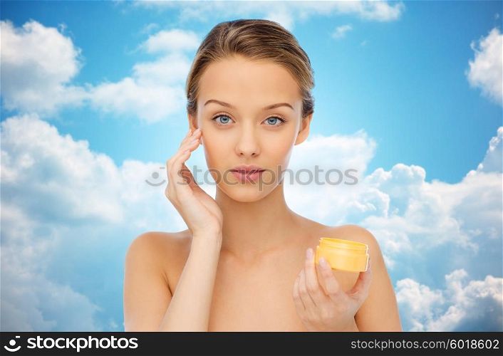 beauty, people, cosmetics, skincare and cosmetics concept - young woman applying cream to her face over blue sky and clouds background