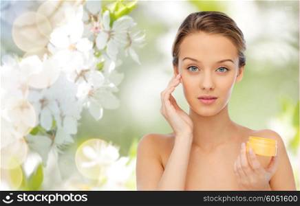 beauty, people, cosmetics, skincare and cosmetics concept - young woman applying cream to her face over green natural background with cherry blossom
