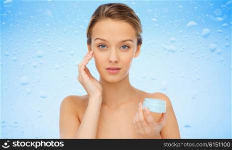 beauty, people, cosmetics, skincare and cosmetics concept - young woman applying cream to her face over water drops on blue background