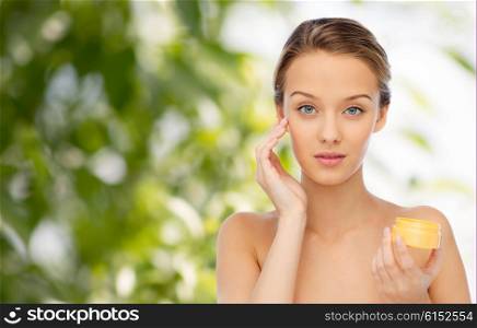 beauty, people, cosmetics, skincare and cosmetics concept - young woman applying cream to her face over green natural background