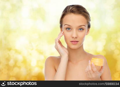 beauty, people, cosmetics, skincare and cosmetics concept - young woman appying cream to her face over yellow holidays lights background