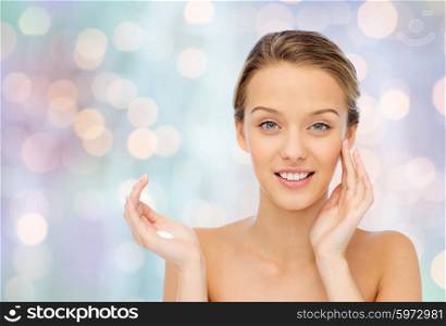 beauty, people, cosmetics, skincare and health concept - happy smiling young woman applying cream to her face over blue holidays lights background