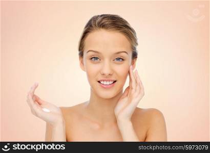 beauty, people, cosmetics, skincare and health concept - happy smiling young woman applying cream to her face over beige background