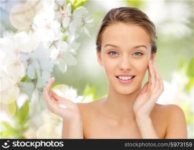 beauty, people, cosmetics, skincare and health concept - happy smiling young woman applying cream to her face over green natural background with cherry blossoms