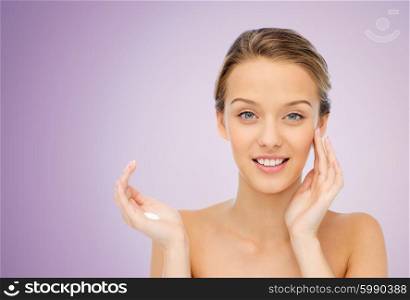 beauty, people, cosmetics, skincare and health concept - happy smiling young woman applying cream to her face over violet background