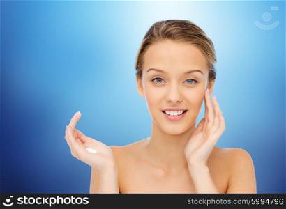 beauty, people, cosmetics, skincare and health concept - happy smiling young woman applying cream to her face over marine blue background