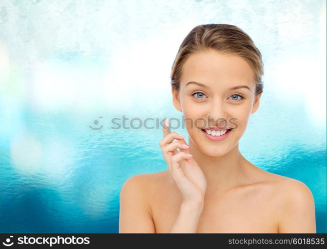 beauty, people, cosmetics, skincare and health concept - happy smiling young woman applying cream to her face over blue water background