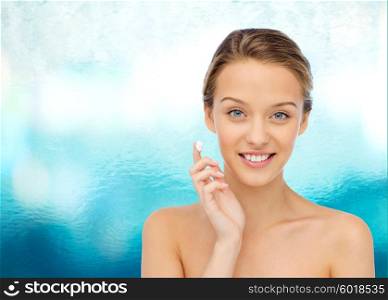 beauty, people, cosmetics, skincare and health concept - happy smiling young woman applying cream to her face over blue water background