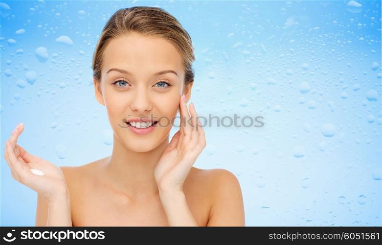 beauty, people, cosmetics, skincare and health concept - happy smiling young woman applying cream to her face over water drops on blue background
