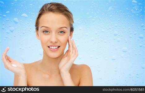beauty, people, cosmetics, skincare and health concept - happy smiling young woman applying cream to her face over water drops on blue background