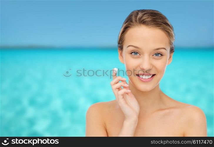 beauty, people, cosmetics, skincare and health concept - happy smiling young woman applying cream to her face over blue sea and sky background