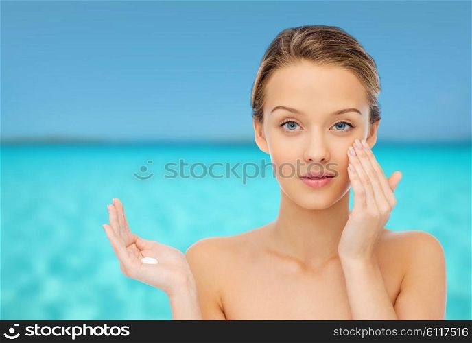 beauty, people, cosmetics, skincare and health concept - happy smiling young woman applying cream to her face over blue sea and sky background