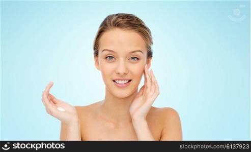 beauty, people, cosmetics, skincare and health concept - happy smiling young woman applying cream to her face over blue background