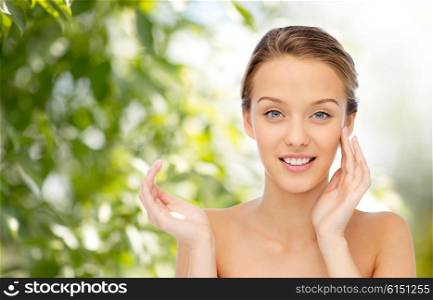 beauty, people, cosmetics, skincare and health concept - happy smiling young woman applying cream to her face over green natural background