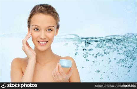 beauty, people, cosmetics, skincare and health concept - smiling young woman applying cream to her face over water splash background