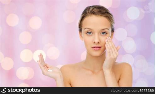beauty, people, cosmetics, skincare and health concept - young woman applying cream to her face over pink lights background