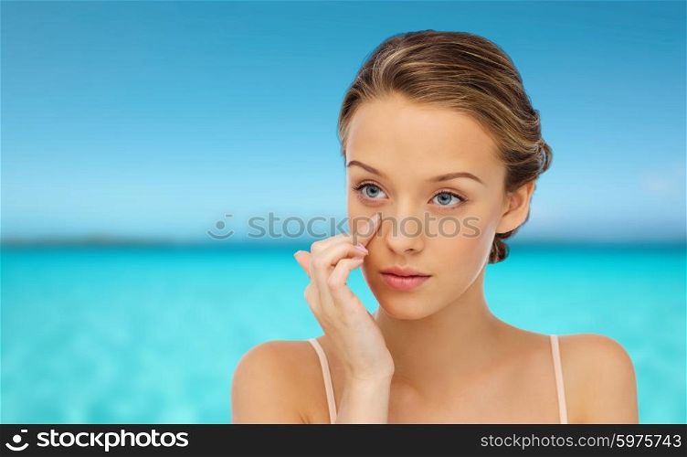beauty, people, cosmetics, skincare and health concept - young woman applying cream to her face over blue sea and sky background