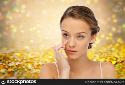 beauty, people, cosmetics, skincare and health concept - young woman applying cream to her face over golden holidays lights or yellow glitter background