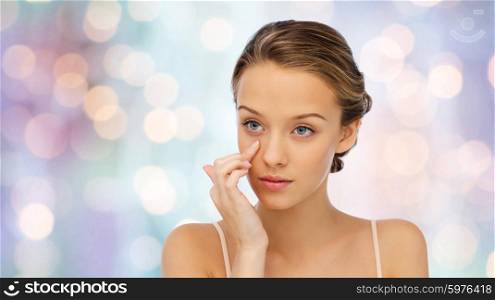 beauty, people, cosmetics, skincare and health concept - young woman applying cream to her face over purple holidays lights background