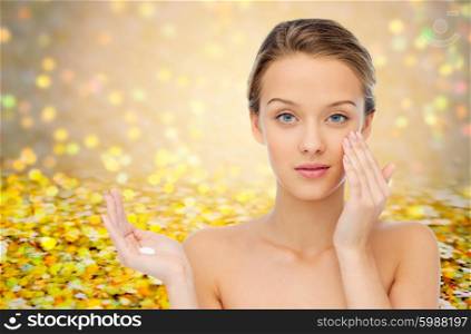 beauty, people, cosmetics, skincare and health concept - young woman applying cream to her face over yellow glitter and confetti background