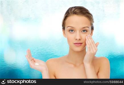 beauty, people, cosmetics, skincare and health concept - young woman applying cream to her face over blue water background