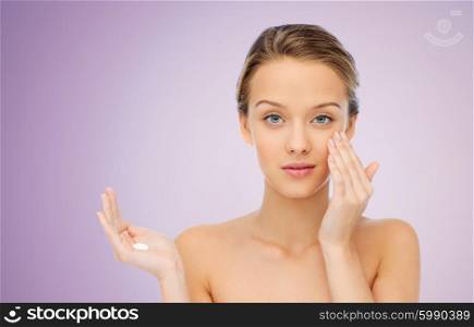 beauty, people, cosmetics, skincare and health concept - young woman applying cream to her face over violet background