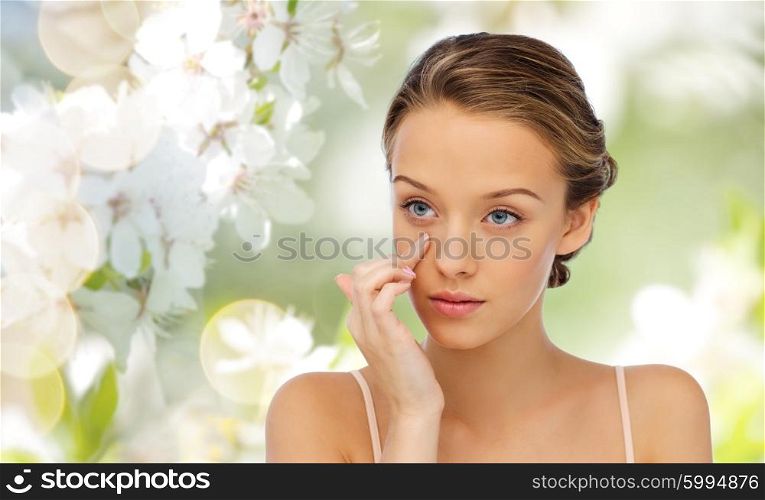 beauty, people, cosmetics, skincare and health concept - young woman applying cream to her face over cherry blossom background