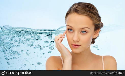 beauty, people, cosmetics, skincare and health concept - young woman applying cream to her face over water splash on blue background