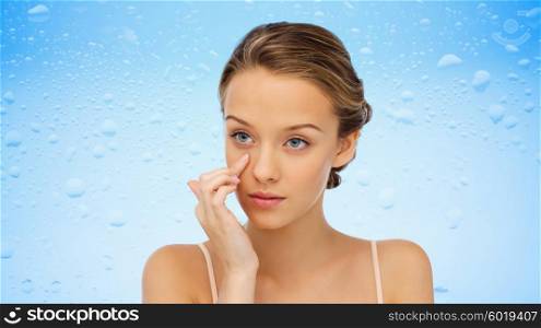 beauty, people, cosmetics, skincare and health concept - young woman applying cream to her face over water drops on blue background