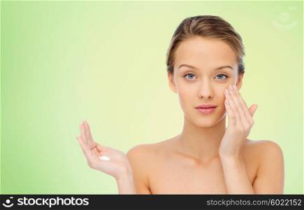 beauty, people, cosmetics, skincare and health concept - young woman applying cream to her face over green natural background