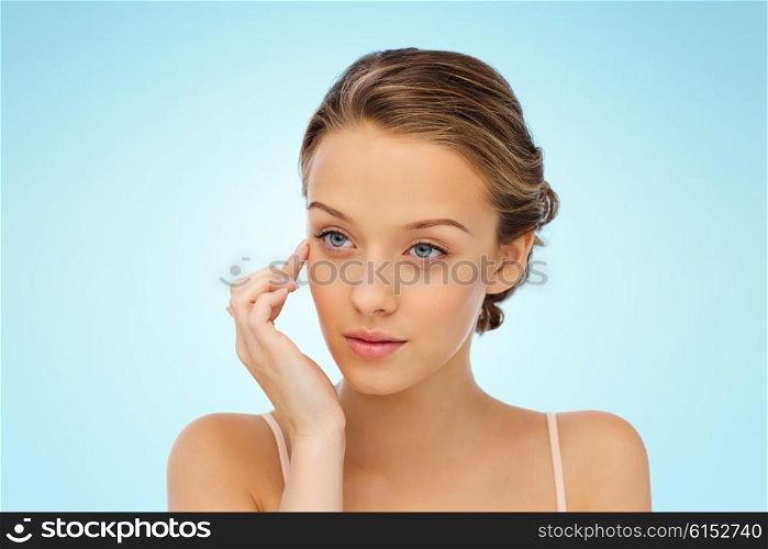 beauty, people, cosmetics, skincare and health concept - young woman applying cream to her face over blue background