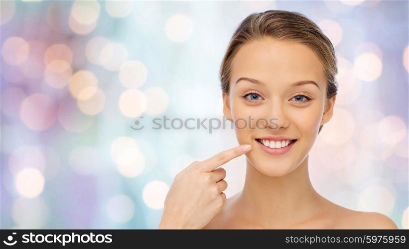 beauty, people, dental care and hygiene concept - happy young woman pointing finger to her smile or teeth over purple holidays lights background
