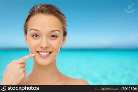 beauty, people, dental care and hygiene concept - happy young woman pointing finger to her smile or teeth over blue sea and sky background