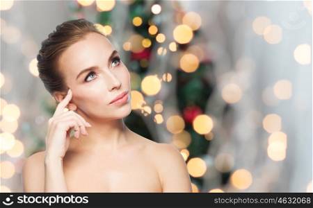 beauty, people, holidays and health concept - beautiful young woman touching her face over christmas tree lights background