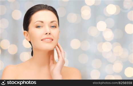 beauty, people, holidays, luxury and health concept - beautiful young woman touching her face over lights background