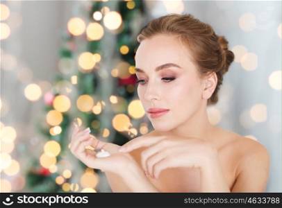 beauty, people, holidays, skincare and cosmetics concept - young woman with moisturizing cream on hand over christmas tree lights background