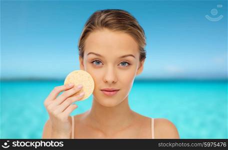 beauty, people, hygiene, and skincare concept - young woman cleaning face with exfoliating sponge over blue sea and sky background