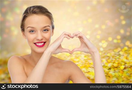 beauty, people, love, valentines day and make up concept - smiling young woman with pink lipstick on lips showing heart shape hand sign over golden glitter or holidays lights background