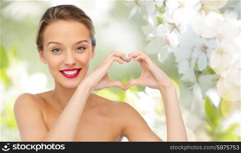 beauty, people, love, valentines day and make up concept - smiling young woman with pink lipstick on lips showing heart shape hand sign over summer green natural background with cherry blossom